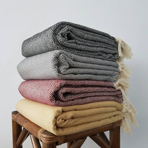 May include: Four woven blankets stacked on a wooden stool. The blankets are in different colors and patterns: black and white, gray and white, red and white, and yellow and white.
