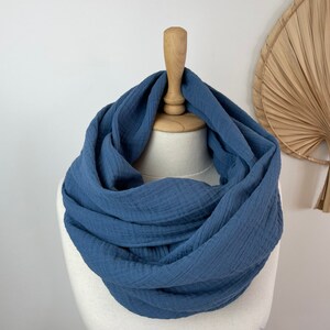 May include: A denim blue infinity scarf made from a textured material, presented on a mannequin. The scarf is styled in a continuous loop, highlighting its soft, flowing form. The backdrop is plain white, with a decorative fan in the background.
