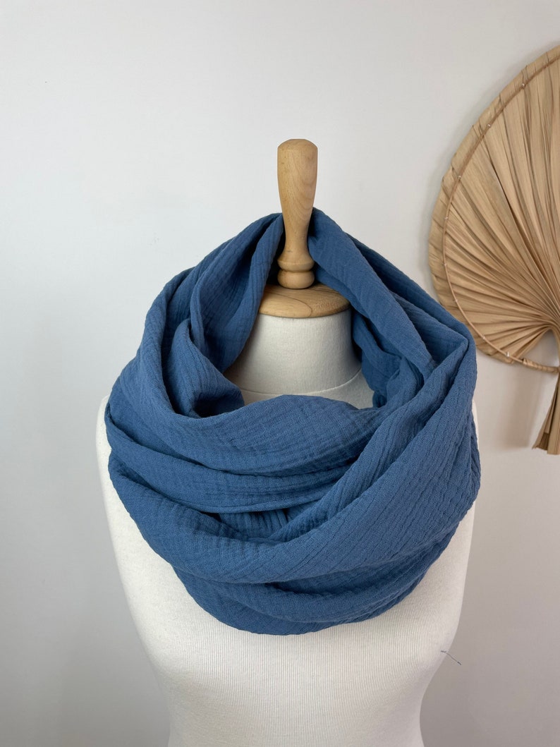 May include: A denim blue infinity scarf made from a textured material, presented on a mannequin. The scarf is styled in a continuous loop, highlighting its soft, flowing form. The backdrop is plain white, with a decorative fan in the background.
