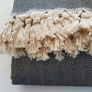 May include: Close-up of a decorative pillow with a black and white herringbone pattern. The pillow features a thick fringe of beige tassels along one edge, adding texture and visual interest. The design is simple yet elegant.