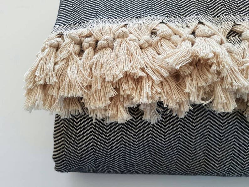May include: Close-up of a decorative pillow with a black and white herringbone pattern. The pillow features a thick fringe of beige tassels along one edge, adding texture and visual interest. The design is simple yet elegant.