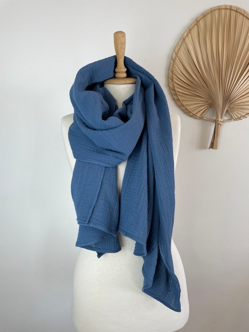 May include: A blue scarf displayed on a mannequin. The scarf is made of a soft, textured material and is draped around the mannequin's neck and shoulders. The scarf's colour is a muted blue, and it has a flowing, lightweight appearance.