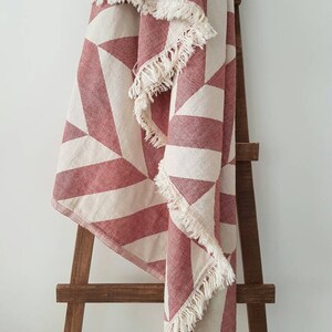 May include: A red and white geometric patterned throw blanket with fringed edges is draped over a wooden ladder. The blanket features a repeating pattern of triangles and stripes in a muted red and off-white color scheme. The fringed edges add a touch of texture.