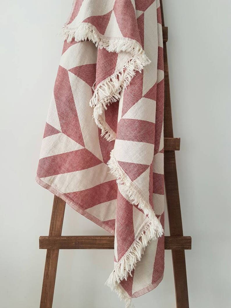 May include: A red and white geometric patterned throw blanket with fringed edges is draped over a wooden ladder. The blanket features a repeating pattern of triangles and stripes in a muted red and off-white color scheme. The fringed edges add a touch of texture.