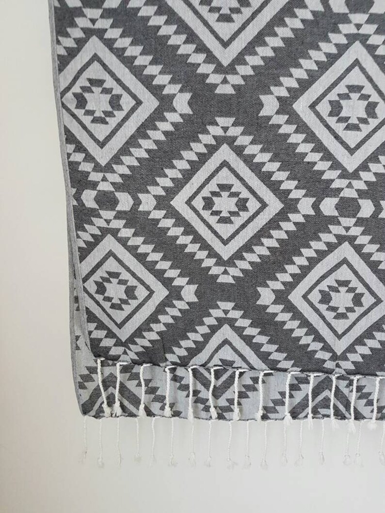May include: A gray and white patterned textile with a geometric design. The pattern features diamond shapes and triangles. The bottom edge has white fringe.