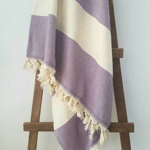 May include: A purple and cream-colored herringbone-patterned blanket with fringe. The blanket is draped over a wooden ladder-style rack. The blanket has wide stripes of cream and purple. The fringe is a cream color.