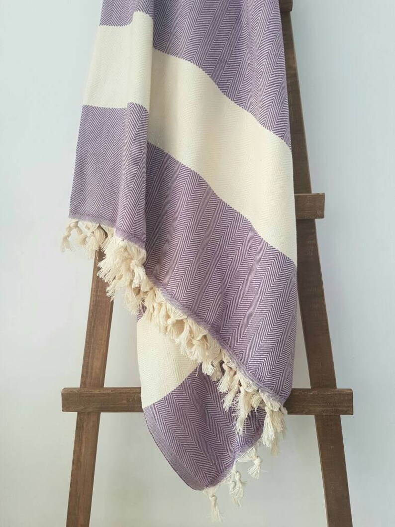 May include: A purple and cream-colored herringbone-patterned blanket with fringe. The blanket is draped over a wooden ladder-style rack. The blanket has wide stripes of cream and purple. The fringe is a cream color.