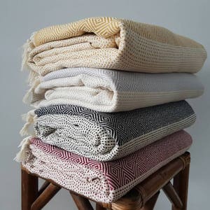May include: A stack of five folded blankets in various colors: yellow, gray, black and white, and burgundy. Each blanket has a geometric diamond pattern and fringe. The blankets are stacked on a small, brown, wooden stool.
