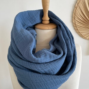 May include: A blue infinity scarf made from a soft, textured material. The scarf is displayed on a mannequin head, highlighting its circular shape. The colour of the scarf is a muted blue, and the fabric looks lightweight.