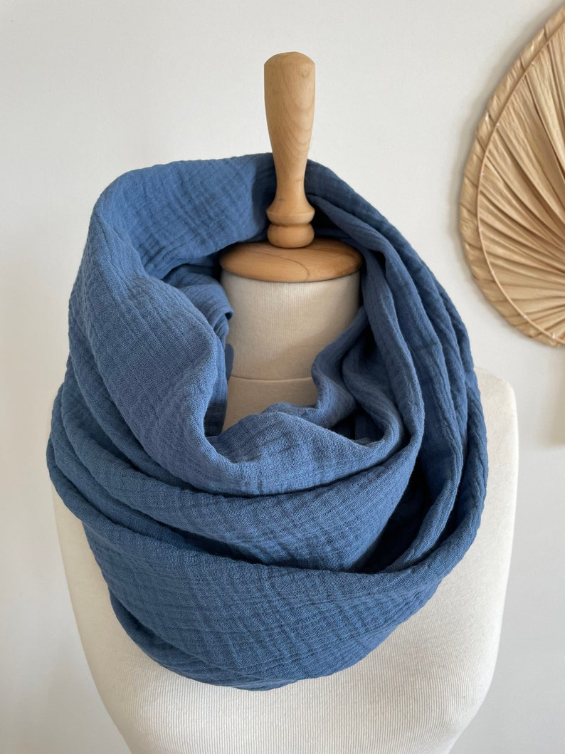 May include: A blue infinity scarf made from a soft, textured material. The scarf is displayed on a mannequin head, highlighting its circular shape. The colour of the scarf is a muted blue, and the fabric looks lightweight.