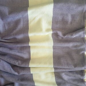 May include: A woven blanket with a herringbone pattern in shades of purple and white. The blanket has a wide white stripe down the center and is finished with cream-colored fringe along the edges. The fabric is soft and appears to be lightweight.