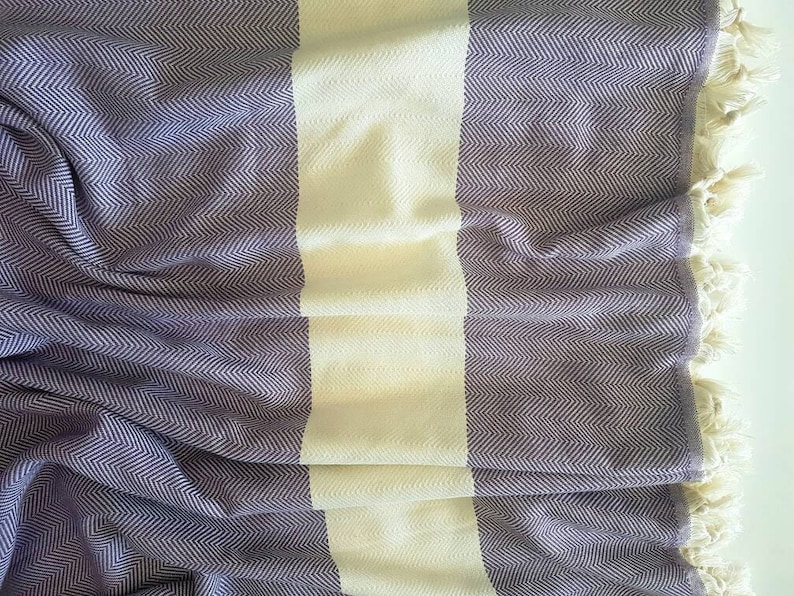 May include: A woven blanket with a herringbone pattern in shades of purple and white. The blanket has a wide white stripe down the center and is finished with cream-colored fringe along the edges. The fabric is soft and appears to be lightweight.