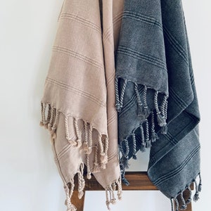 May include: Two striped towels with fringe, one in a light beige color and the other in a dark gray color, hanging on a wooden ladder.