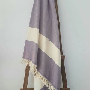 May include: A lavender and cream-colored woven throw blanket with a herringbone pattern and cream-colored fringe. The blanket is draped over a wooden ladder, showcasing its design and texture. The blanket has a cream-colored stripe.