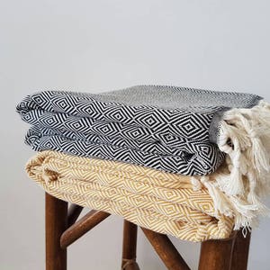 May include: Three folded blankets with geometric diamond patterns are stacked on a wooden stool. The top blanket is gray, the middle one is black and white, and the bottom one is yellow. The black and white blanket has white fringe.