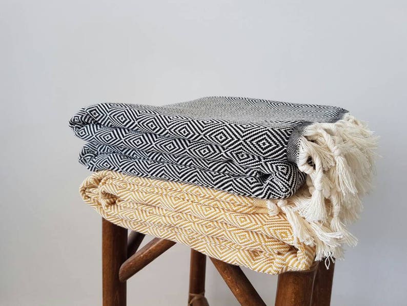 May include: Three folded blankets with geometric diamond patterns are stacked on a wooden stool. The top blanket is gray, the middle one is black and white, and the bottom one is yellow. The black and white blanket has white fringe.