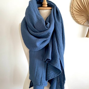 May include: A blue scarf displayed on a mannequin. The scarf is made of a soft, textured material and is draped in flowing folds. The colour of the scarf is a muted, medium blue, and it appears to be lightweight. The background is a plain white wall.