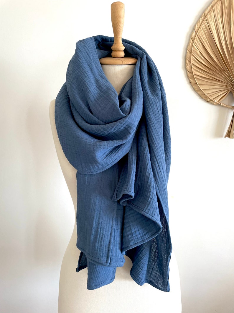 May include: A blue scarf displayed on a mannequin. The scarf is made of a soft, textured material and is draped in flowing folds. The colour of the scarf is a muted, medium blue, and it appears to be lightweight. The background is a plain white wall.