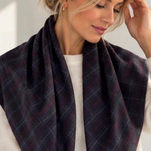 May include: A dark charcoal gray scarf featuring a subtle plaid pattern with thin intersecting lines in shades of deep burgundy and muted white. The scarf is draped around the neck over a cream-colored knit sweater and blue denim jeans.