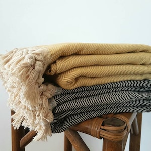 May include: Two folded blankets, one mustard yellow and one black and white herringbone pattern, are stacked on a wooden stool. The top blanket has a fringe of cream-colored tassels. The blankets appear soft and cozy.