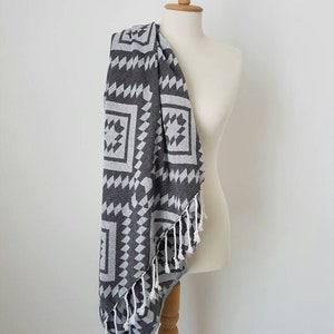 May include: A dark gray and white patterned towel with a geometric design is draped over a mannequin. The towel has a fringe along one edge. The pattern includes squares and triangles. The towel is made of a soft material.