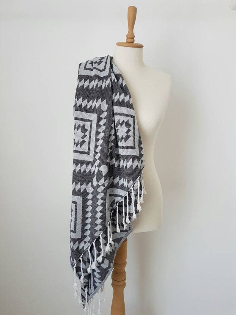 May include: A dark gray and white patterned towel with a geometric design is draped over a mannequin. The towel has a fringe along one edge. The pattern includes squares and triangles. The towel is made of a soft material.