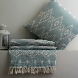 May include: A light blue and white patterned pillow and folded blanket with a fringed edge. A matching pillow is propped up against a white wall. A white lantern sits to the left of the blanket.
