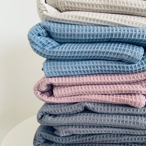 Peut inclure: Une pile de couvertures en tissu gaufré de différentes couleurs, dont le beige, le bleu, le rose et le gris.