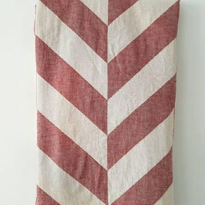 May include: A red and white chevron patterned blanket with a fringed edge. The blanket is folded, displaying a repeating V-shaped design. The color scheme is a warm red and off-white, creating a cozy aesthetic. This blanket is suitable for home decor.