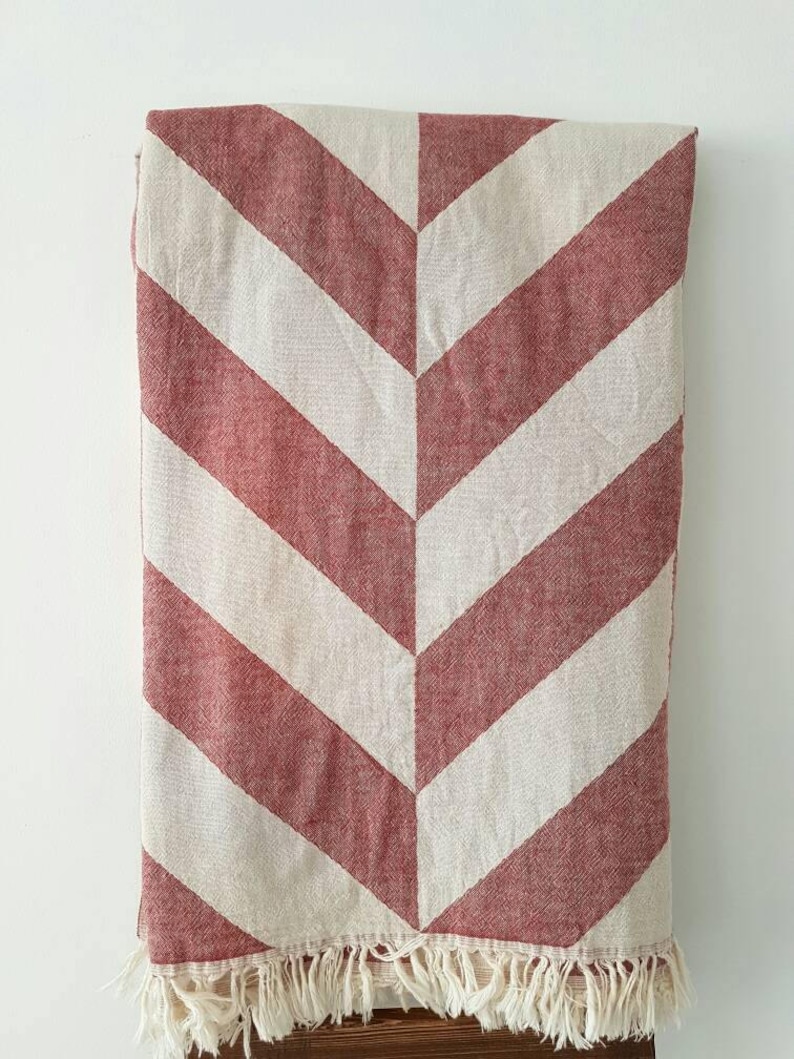 May include: A red and white chevron patterned blanket with a fringed edge. The blanket is folded, displaying a repeating V-shaped design. The color scheme is a warm red and off-white, creating a cozy aesthetic. This blanket is suitable for home decor.