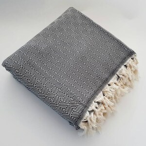 May include: Folded black and white patterned blanket with a diamond design. The blanket has a gray border and cream-colored fringe along one edge. The blanket is folded neatly on a white surface.