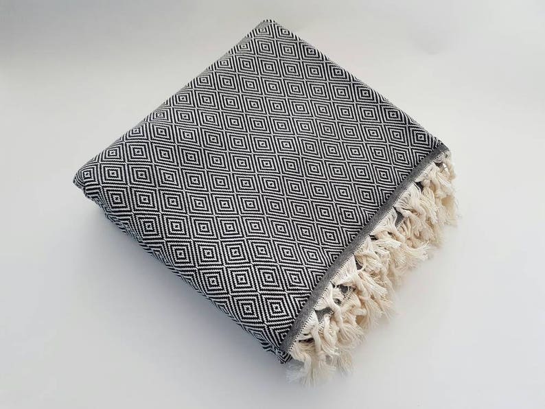 May include: Folded black and white patterned blanket with a diamond design. The blanket has a gray border and cream-colored fringe along one edge. The blanket is folded neatly on a white surface.