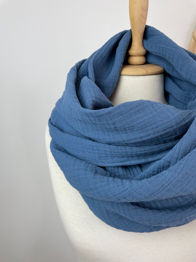 May include: A blue infinity scarf made from a soft, textured material. The scarf is displayed on a mannequin, highlighting its circular design and how it would be worn. The colour is a muted, dusty blue.
