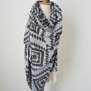 May include: A gray and black patterned scarf with a geometric design, displayed on a mannequin. The scarf features a repeating pattern of triangles and diamonds. White fringe accents the ends of the scarf. The scarf is made of a soft, lightweight material.
