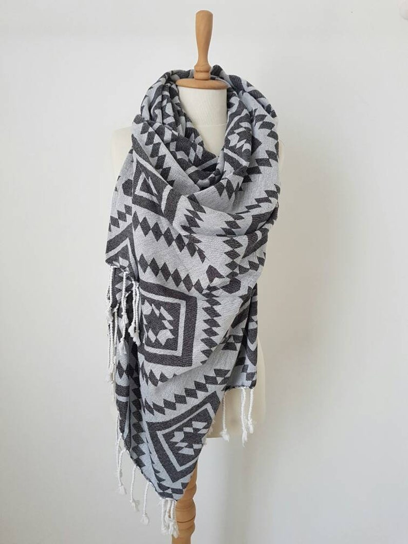 May include: A gray and black patterned scarf with a geometric design, displayed on a mannequin. The scarf features a repeating pattern of triangles and diamonds. White fringe accents the ends of the scarf. The scarf is made of a soft, lightweight material.