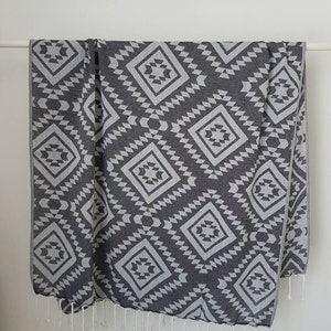 May include: A dark gray and white patterned textile, possibly a blanket or towel, hangs against a white background. The design features repeating diamond shapes with geometric patterns. White fringe trims the bottom edge.