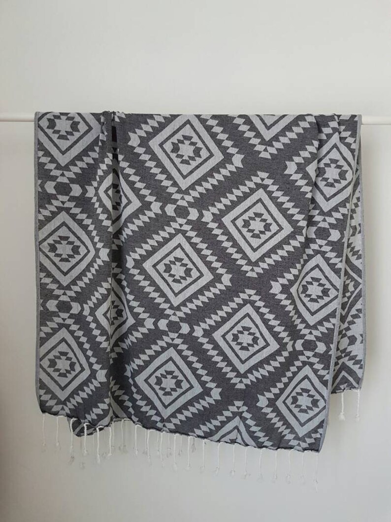 May include: A dark gray and white patterned textile, possibly a blanket or towel, hangs against a white background. The design features repeating diamond shapes with geometric patterns. White fringe trims the bottom edge.