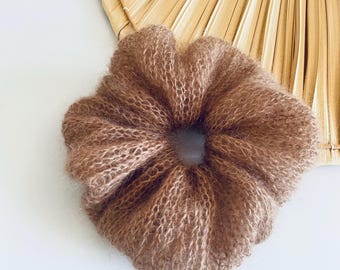 May include: A brown, fluffy scrunchie made of a soft, textured material. The scrunchie is round with a hole in the center. A dried palm leaf fan is in the background.