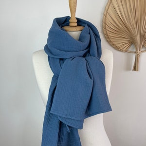 May include: A blue scarf displayed on a mannequin. The scarf, in a solid blue hue, is made from a soft, textured material. It's draped around the mannequin's neck and shoulders, with the ends falling down. The background is a plain white.