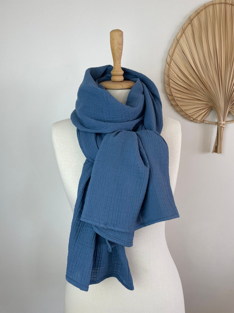 May include: A blue scarf displayed on a mannequin. The scarf, in a solid blue hue, is made from a soft, textured material. It's draped around the mannequin's neck and shoulders, with the ends falling down. The background is a plain white.