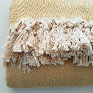 May include: A folded yellow blanket with a herringbone pattern and a cream-colored fringe. The fringe is made of knotted tassels. The blanket is a warm, neutral color, suitable for home decor.