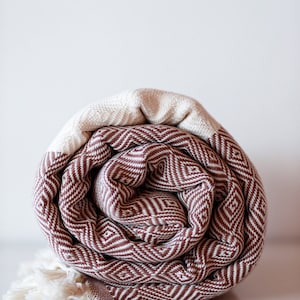 May include: A rolled up brown and white patterned Turkish towel with fringe. The towel is made of cotton and has a geometric design.