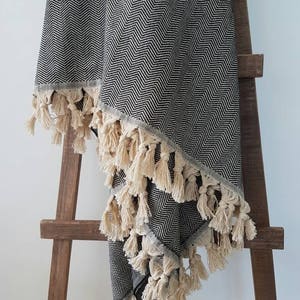 May include: A black and white herringbone patterned throw blanket with cream-colored tassels. The blanket is draped over a wooden ladder, showcasing its texture and design. The tassels are evenly spaced along the edges, adding a decorative element.