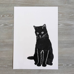 May include: A black and white linocut print of a seated cat. The cat has a solid black body with white details in its eyes and whiskers. The print is on white paper, with a simple, minimalist style.