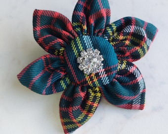 May include: A fabric flower brooch featuring a dark green, red, and yellow plaid tartan pattern. The flower has five rounded petals radiating from a central fabric-covered button, which is accented with a clear, faceted, star-shaped crystal.