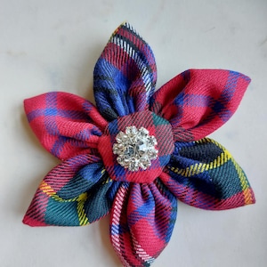 May include: A fabric flower in a tartan pattern of red, blue, green, and yellow. The flower has six petals and a central, round, red fabric piece adorned with a clear, sparkling rhinestone.