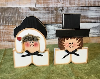 May include: Wooden bride and groom figurines. The bride has a black hat, a red heart, and a black bow tie. The groom has a black top hat and a black bow tie. The figurines are painted with rosy cheeks and smiling faces.