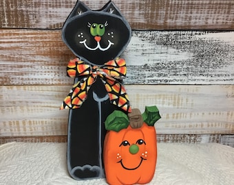 May include: Black cat with a candy corn bow and a smiling orange pumpkin with green leaves and a brown stem.  The cat and pumpkin are wooden and hand-painted.