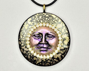 Puede incluir: Un collar con colgante redondo con un diseño de cara de sol. El colgante tiene un borde negro con detalles dorados y una cara de sol central en tonos rosas y morados. El collar tiene un cordón negro.