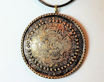 Puede incluir: Un collar con un colgante redondo adornado con diseños intrincados. El colgante es de color dorado y está unido a un cordón de cuero negro.